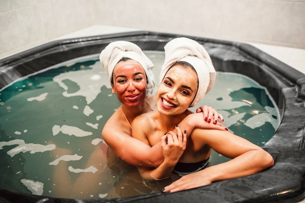 Cheerful and positive young women look   and embrace each other. They sit in hydromassage bath full of water. Women are happy. They have beauty masks on face.