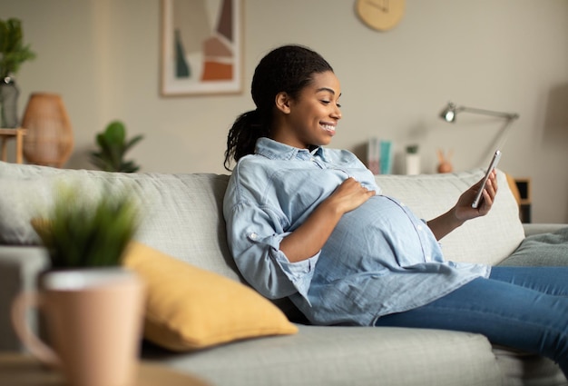 Photo cheerful pregnant african lady using smartphone video calling at home