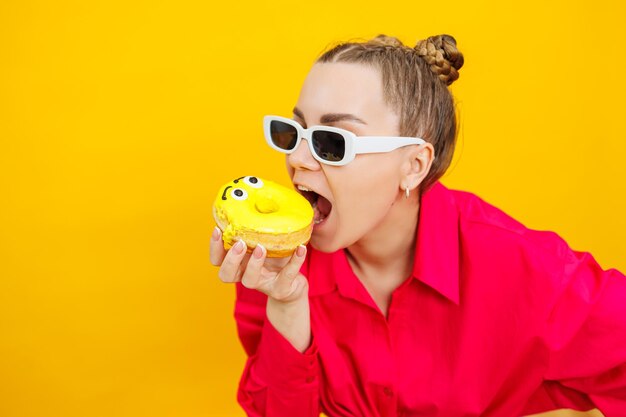 Cheerful pregnant woman holding a sweet yellow donut in her hand on a yellow background expecting a child pregnancy and motherhood the concept of healthy and unhealthy food diet junk food