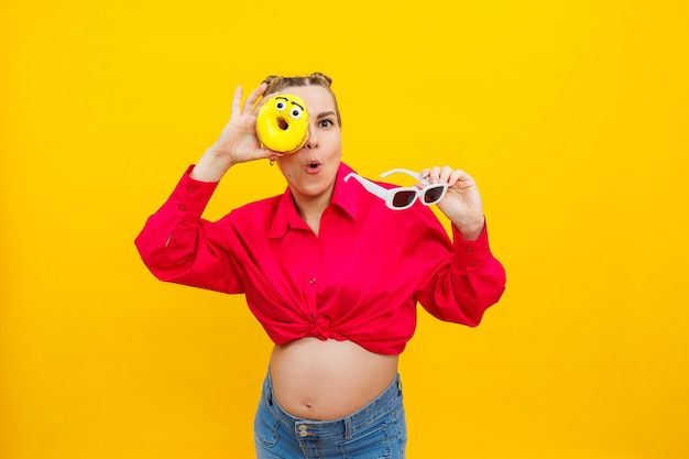 黄色の背景に甘い黄色のドーナツを手に持った陽気な妊婦子供の妊娠と母性を期待健康と不健康な食品ダイエットの概念ジャンク フード