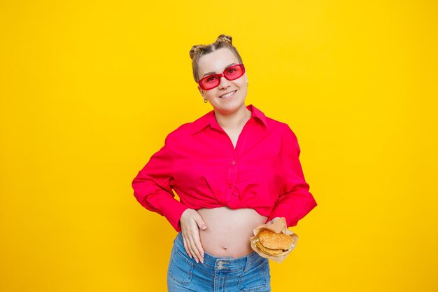 Foto donna incinta allegra in camicia rosa che tiene hamburger su sfondo giallo isolato con espressione facciale sorpresa e scioccata cibo dannoso durante la gravidanza una donna incinta mangia fast food