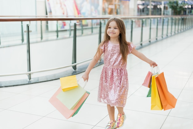 쇼핑백과 함께 걷는 명랑 유치원 소녀. 쇼핑 가방가 게에서 포즈와 함께 꽤 웃는 소녀. 상점에서의 쇼핑 개념