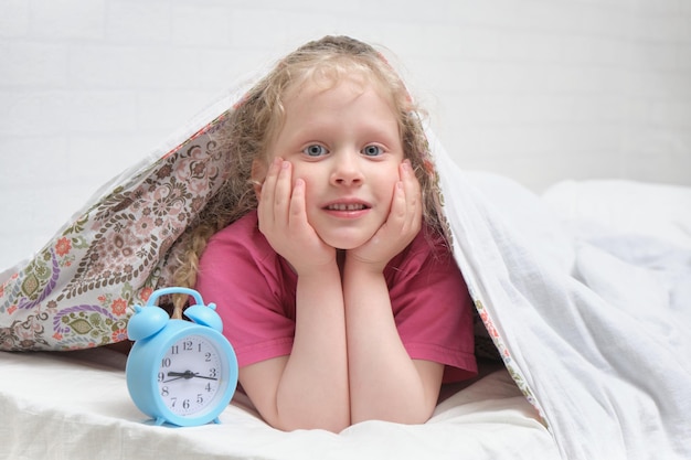A cheerful preschooler girl in a pink tshirt lies under the blanket and next to the alarm clock