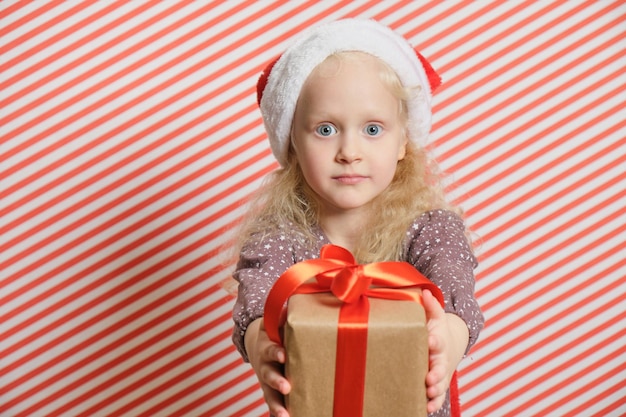 赤い縞模様の背景にギフト、ブロンドの髪の少女の手に赤いリボンのギフト、ボクシングデー、クリスマスプレゼントのコンセプトとサンタの帽子をかぶった陽気な未就学児の女の子