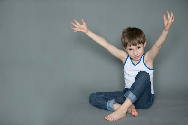 Cheerful preschooler sitting on the floor hands up Smiling boy in jeans