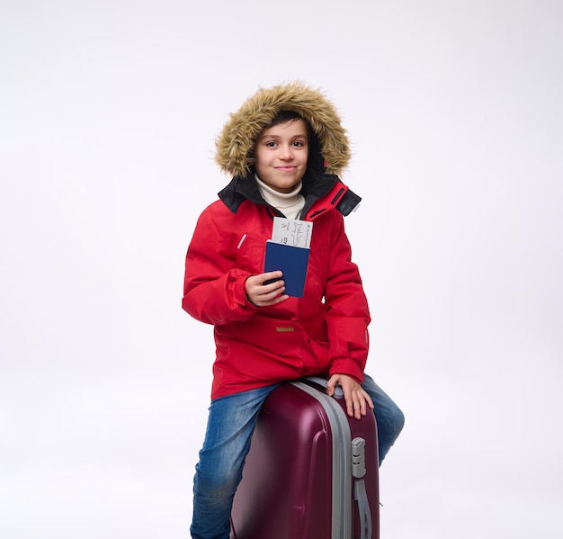 A cheerful preteen, elementary aged boy in bright red down jacket sitting on his luggage, looking at camera and showing passport with boarding pass. Winter holidays, travel concept with copy ad space