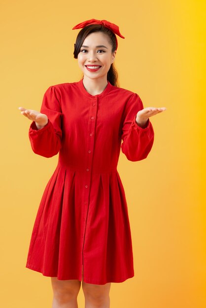 Cheerful pretty Asian woman in red dress showing two brank hands over orange background