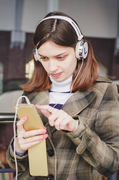 Cheerful pretty brunette girl in a shopping center having fun listening to music in headphones and selecting music from her phone