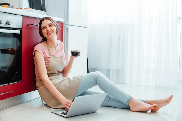 Cheerful pretty lady sitting by the cooker on a kitchen floor and smiling while touching the laptop keyboard