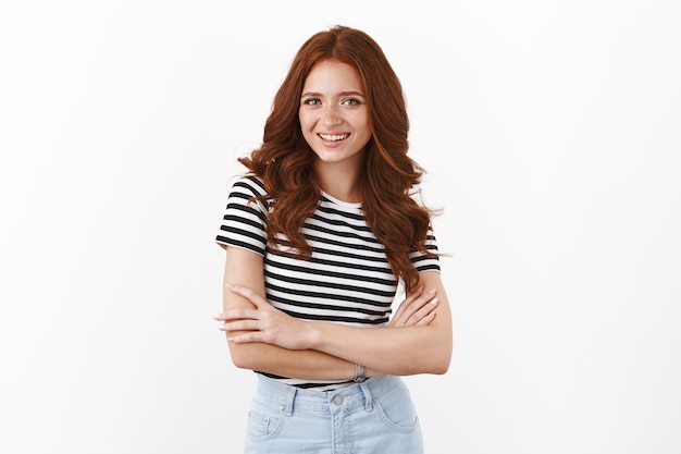 Cheerful pretty redhead girl in striped t-shirt, cross arms and smiling confident, looking determined and self-assured, standing white wall