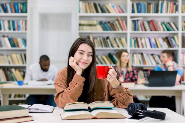 カジュアルな服装で陽気なかなりスマートな女の子の学生、カメラ目線と笑顔、一杯のコーヒーまたは紅茶を保持し、大学図書館での試験の準備