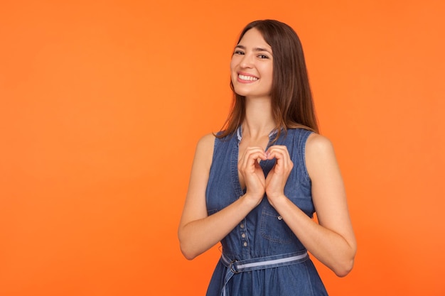 Cheerful pretty woman in dress making heart shape with hands and smiling kindly showing charity symbol declaration of love expressing emotions indoor studio shot isolated on orange background