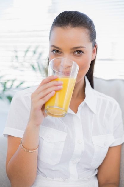 Cheerful pretty woman drinking orange juice sitting on cosy couch