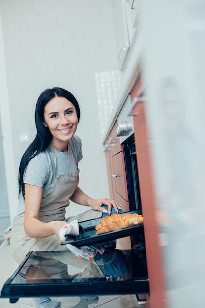 Cheerful pretty woman in the kitchen holding a hot baking pan with delicious croissants