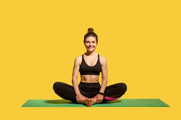 Cheerful pretty woman with hair bun in tight sportswear sitting in lotus position on gym mat practicing yoga doing sports and smiling to camera happy healthy lifestyle indoor studio shot isolated