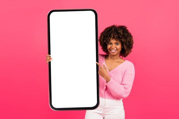 Веселая симпатичная молодая афро-американская кудрявая женщина в повседневной одежде с брекетами держит большой смартфон с пустым экраном