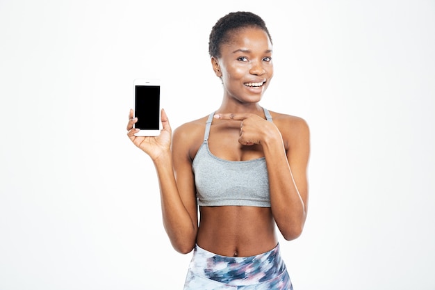 Cheerful pretty young african american sportswoman holding blank screen smartphone and pointing on it over white background