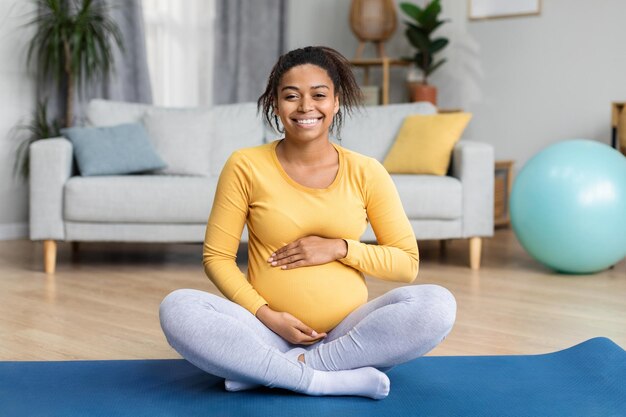 Cheerful pretty young black pregnant lady with big belly\
practicing yoga on mat in modern living room interior