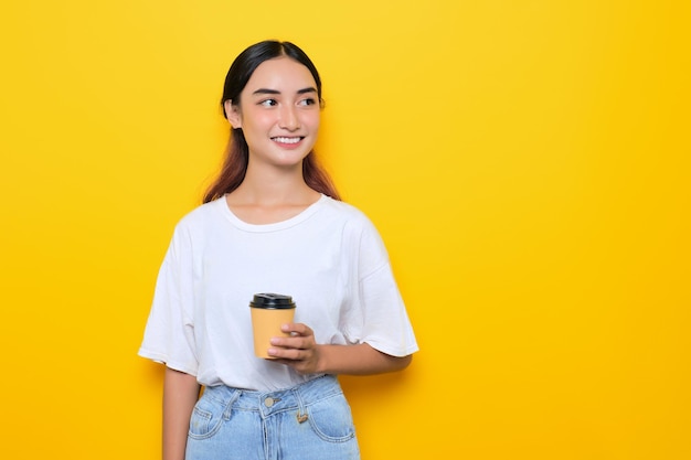 Cheerful pretty young girl in white tshirt holding coffee cup and looking away at blank space isolated on yellow background