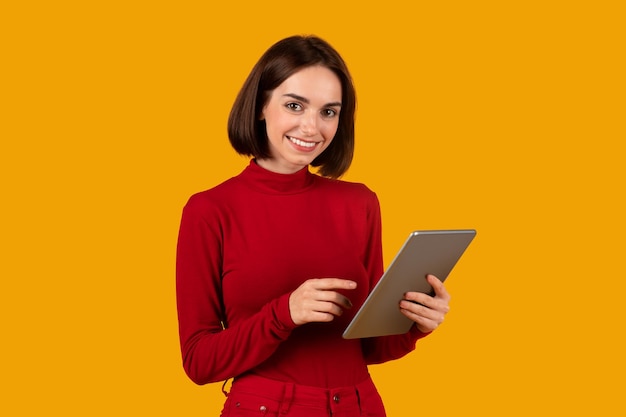 Веселая симпатичная молодая женщина цифровой кочевник с помощью планшета на оранжевом