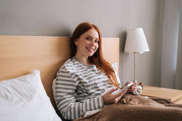 Cheerful pretty young woman shopping online with credit card and smartphone sitting in bed at home looking at camera