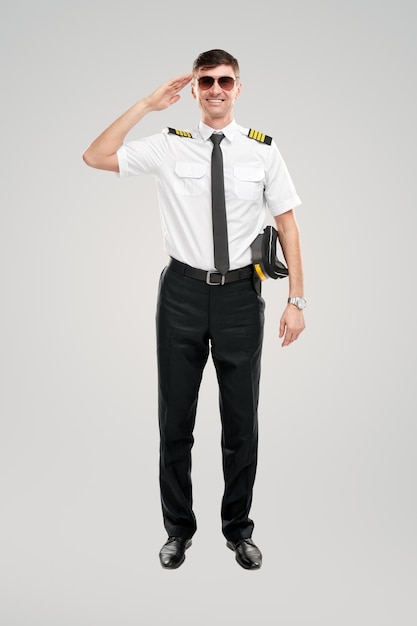 Cheerful professional male airline captain in uniform saluting while standing on gray background in studio and looking at camera