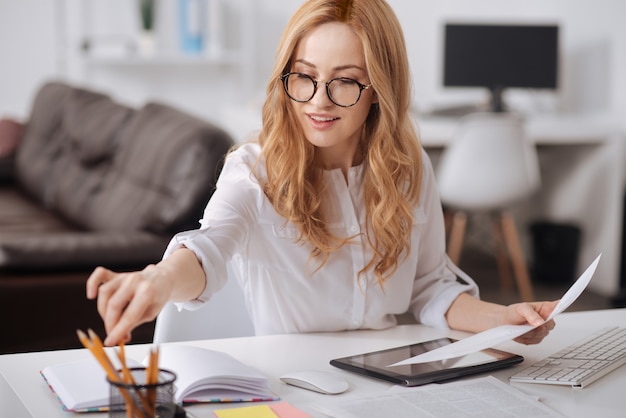 Cheerful qualified young manager sitting at the table at the workplace and working while using tablet and making notes