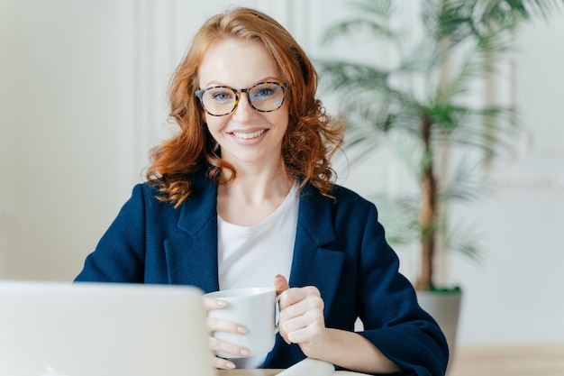 Cheerful redhead woman prepares application letter sits in
front of opened laptop computer browses internet and seeks for job
vacancies holds mug with hot drink wears spectacles and formal
outfit