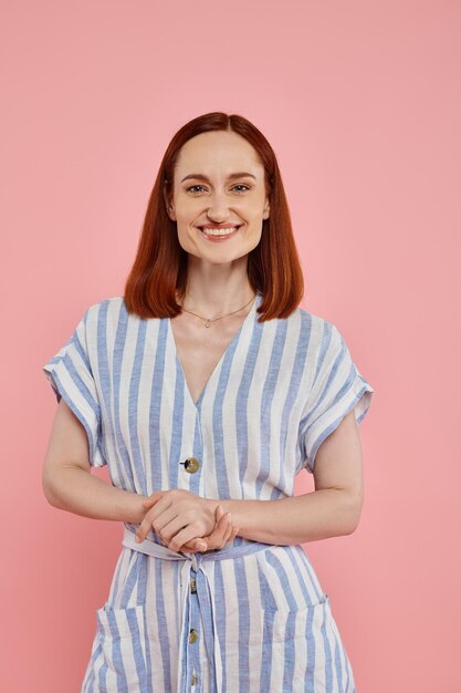 cheerful redhead woman in striped dress looking at camera on pink backdrop studio portrait