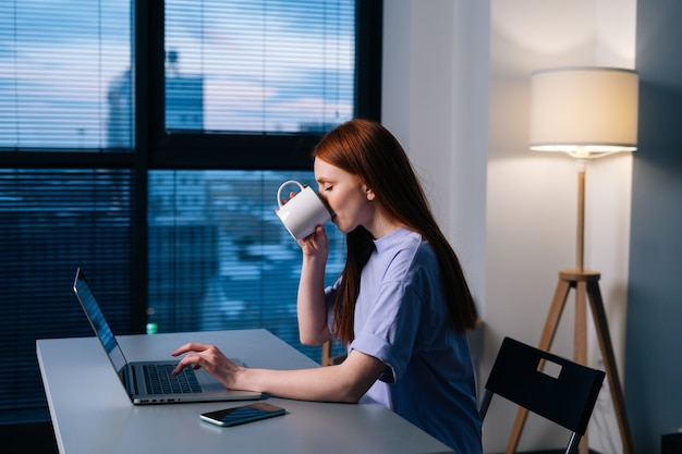 暗いオフィスの机に座って、マグカップからコーヒーを飲んでラップトップで入力して陽気な赤毛の若いビジネス女性。夜遅くに窓の近くの机に座ってコンピューターに取り組んでいる幸せな実業家。