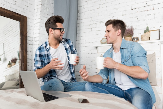 Cheerful relaxed homosexual couple talking and having a drink while sitting together on the bed.