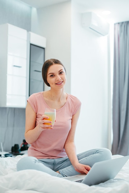 Cheerful relaxed woman enjoying tasty beverage in the bedroom while sitting with crossed legs on the bed