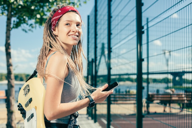 Cheerful relaxed young lady with dreadlocks smiling and looking happy while being near the sports ground with her guitar