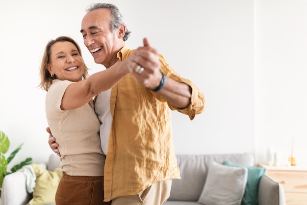 Cheerful retired spouses husband and wife dancing and laughing in living room happy romantic couple enjoying slow dance