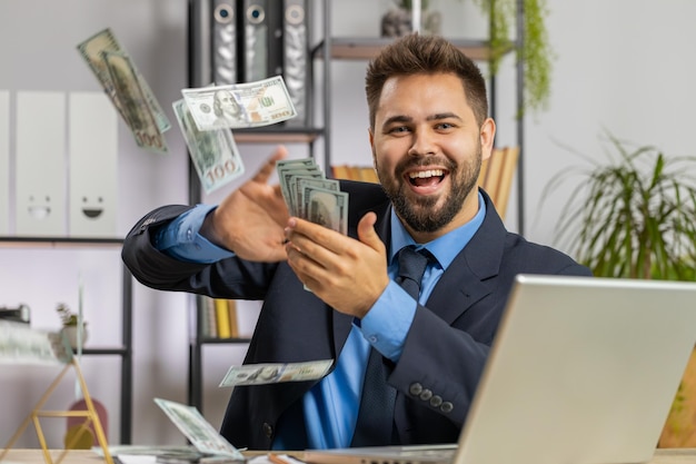 Веселый богатый бизнесмен, работающий на офисном ноутбуке, тратящий деньги на камеру, получает прибыль.