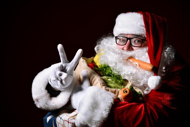 Cheerful Santa Claus is holding a big bag full of fruits and vegetables Shows shows peace sign