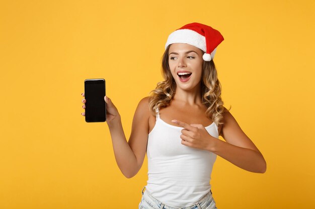 Cheerful santa girl in christmas hat posing isolated on yellow background. happy new year 2020 celebration holiday concept. mock up copy space. pointing finger on mobile phone with blank empty screen