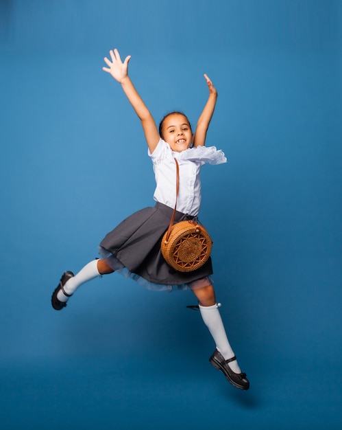 Cheerful schoolgirl 7 years old with a bag over her shoulder, jumping on a blue background.