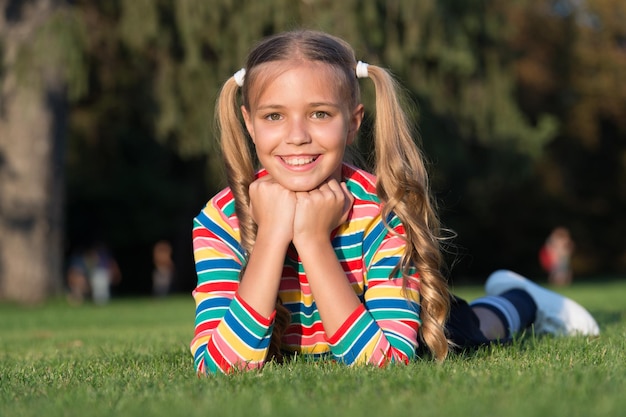 Cheerful schoolgirl on sunny day Girl ponytails hairstyle enjoy relax Living happy life Happy smiling pupil Have fun Girl kid laying green grass Healthy emotional happy kid relaxing outdoors