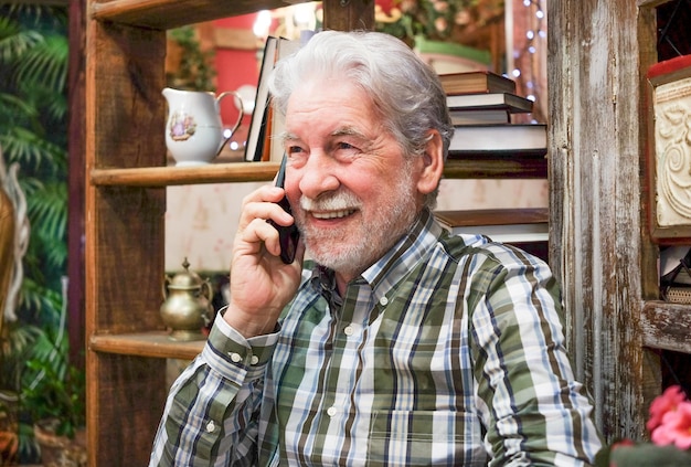 Photo cheerful senior 70s grandfather sit at home talking on modern cellphone, happy mature 70s man relax in living room speaks having pleasant smartphone call