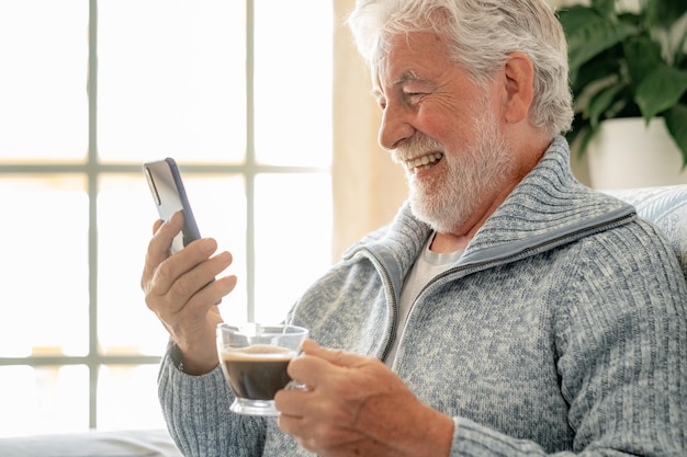 Cheerful senior bearded man chatting on mobile phone while seated on sofa with a coffee cup Elderly male using cellphone enjoying leisure and relax