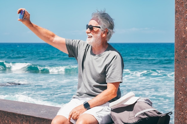 Cheerful Senior Bearded Man Sitting Close the Beach at Sea Making Selfie by Smartphone