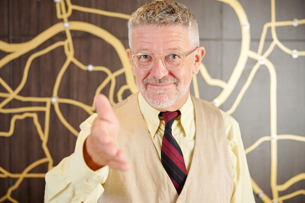 Cheerful senior businessman in glasses outstretching hand for handshake and greeting partner