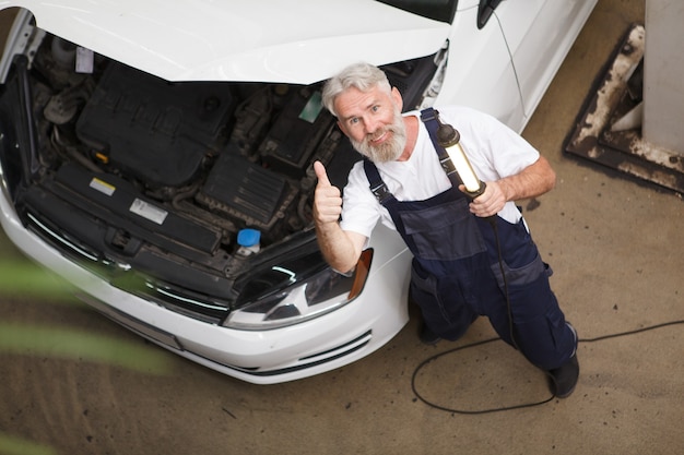 Cheerful senior car mechanic smiling showing thumbs up while repairing an automobile at the garage