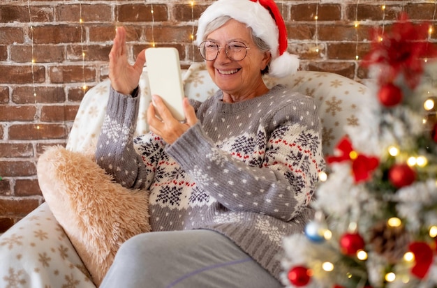 Cheerful senior caucasian woman in winter sweater and Santa hat sitting alone at home using phone video chatting with family or friends celebrate Christmas and new year