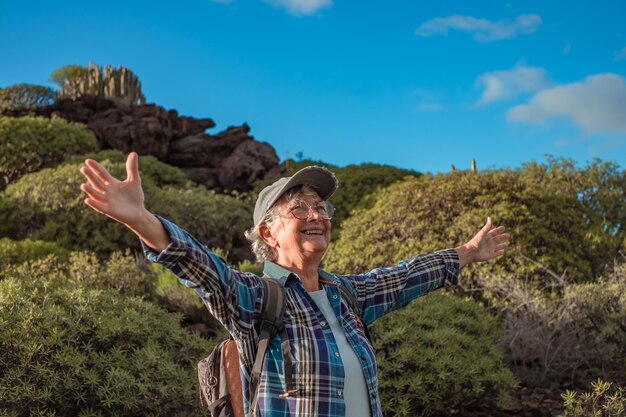 Cheerful senior caucasian woman with open arms in mountain trekking enjoying nature and freedom Smiling mature lady in hat and casual clothes among green bushes and blue sky