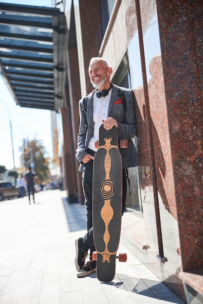 Cheerful senior citizen smiling and standing next to a wall while holding his longboard