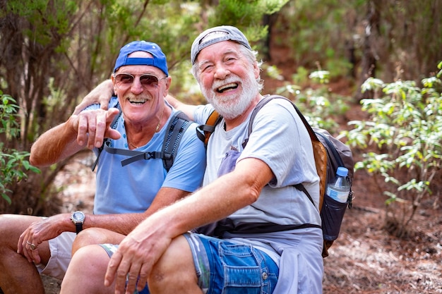 Cheerful senior couple of friends in mountain excursion looking at camera laughing while sitting to rest enjoying healthy lifestyle in nature