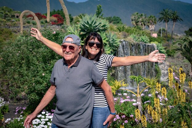 Веселая пожилая пара в солнечных очках наслаждается солнечным днем в красивом саду с водопадом