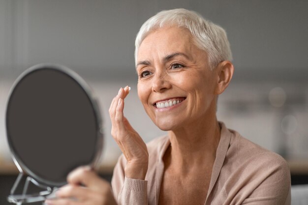 Photo cheerful senior lady applying facial moisturizer looking at mirror indoor